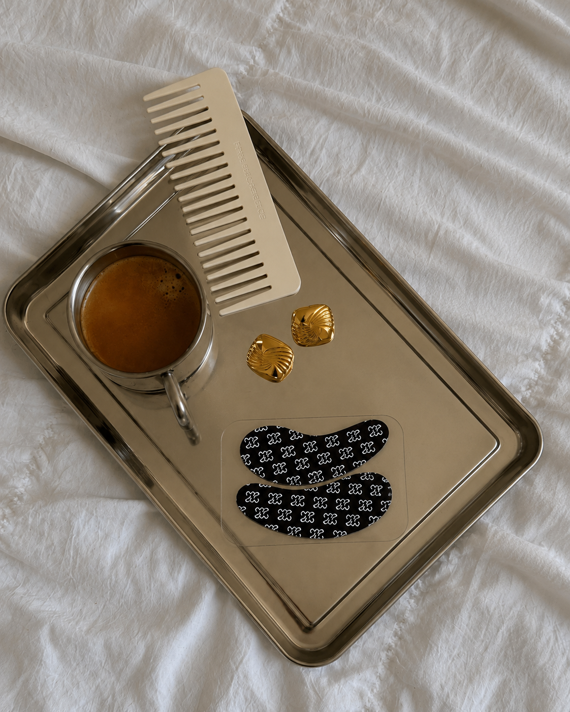 Every Morning Eye Mask by The Morning Pause styled on a silver tray with espresso, gold earrings, and a wide-tooth comb on white linen